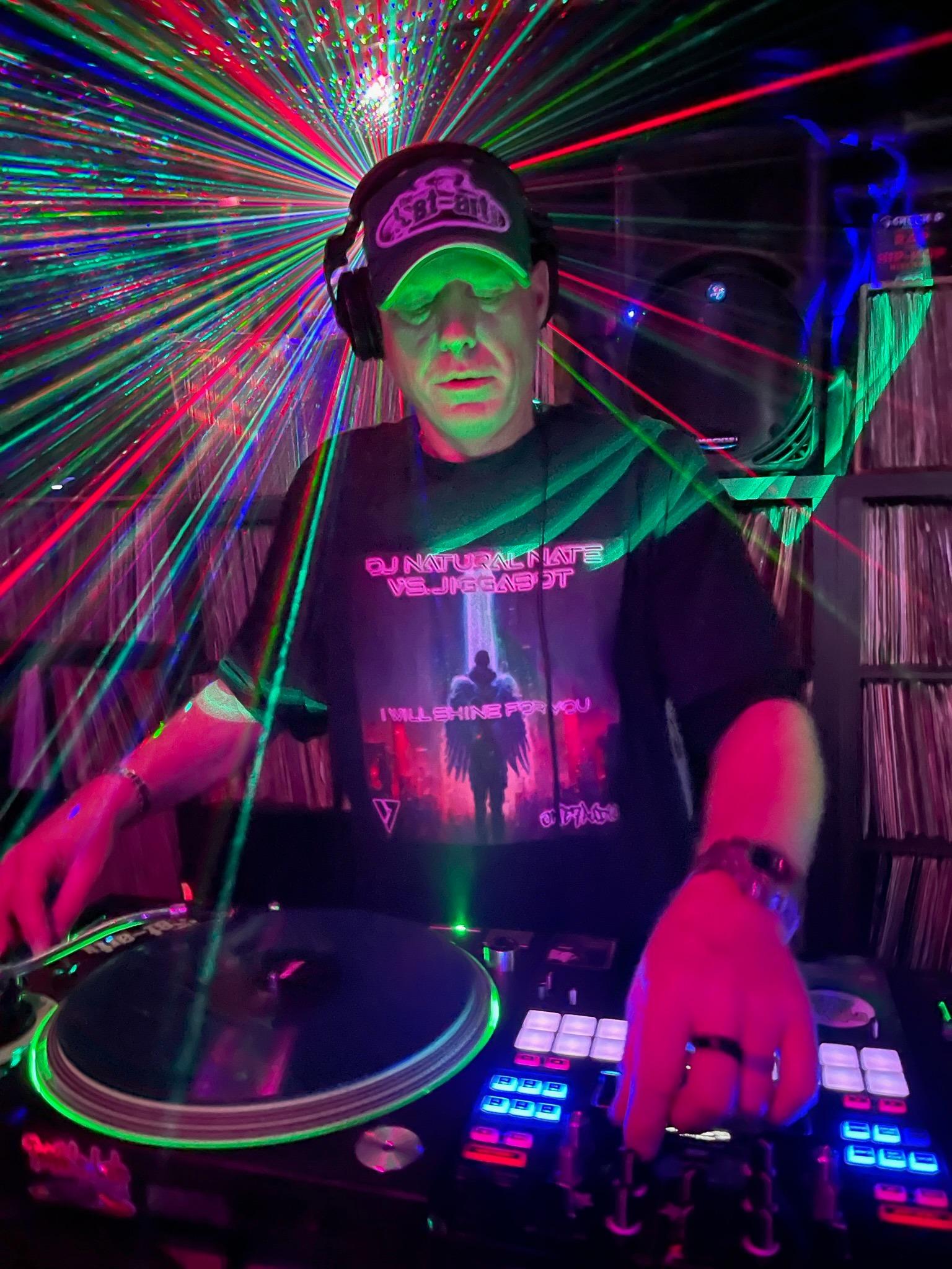 DJ Natural Nate® in "I Will Shine For You" shirt under full rainbow laser explosion with Pioneer DJ pads and TLA hat